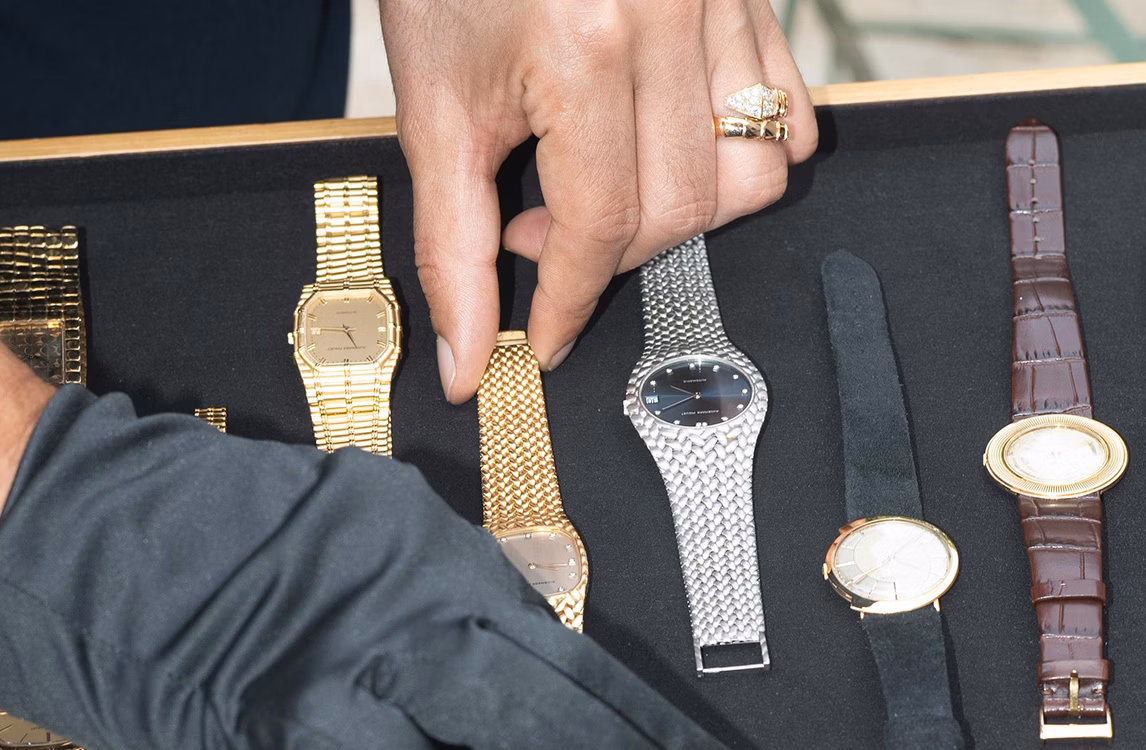 Hand with rings selecting an Audemars Piguet Mini Bamboo watch from a tray of vintage watches, including a gold octagonal model, a silver mesh watch, and the reference 5182 watch with a black strap.