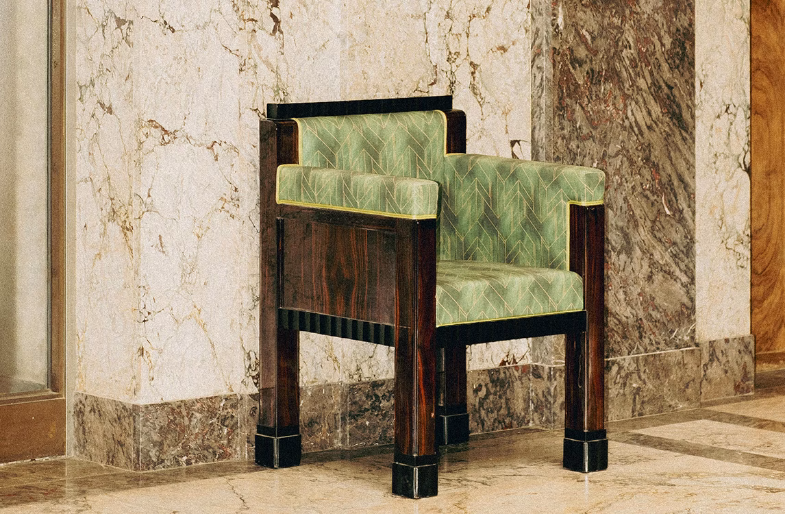 Art Deco armchair with a dark wood frame and green geometric upholstery against a marble wall.