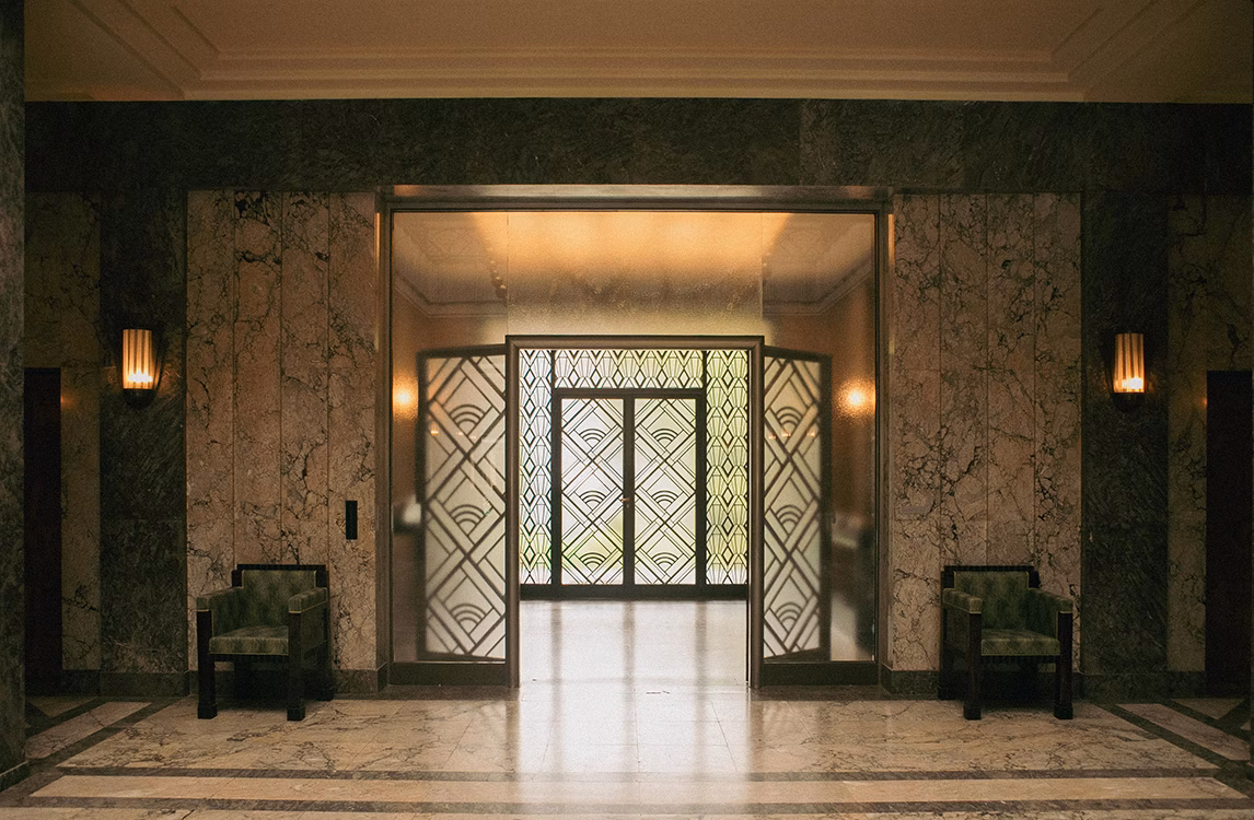 Art Deco lobby entrance at Villa Empain, with marble walls, geometric glass doors, and two green chairs.