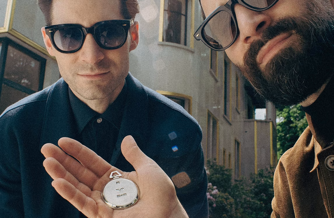 Ryan and Adam Goldston outdoors in sunglasses, one holding an open silver pocket watch.