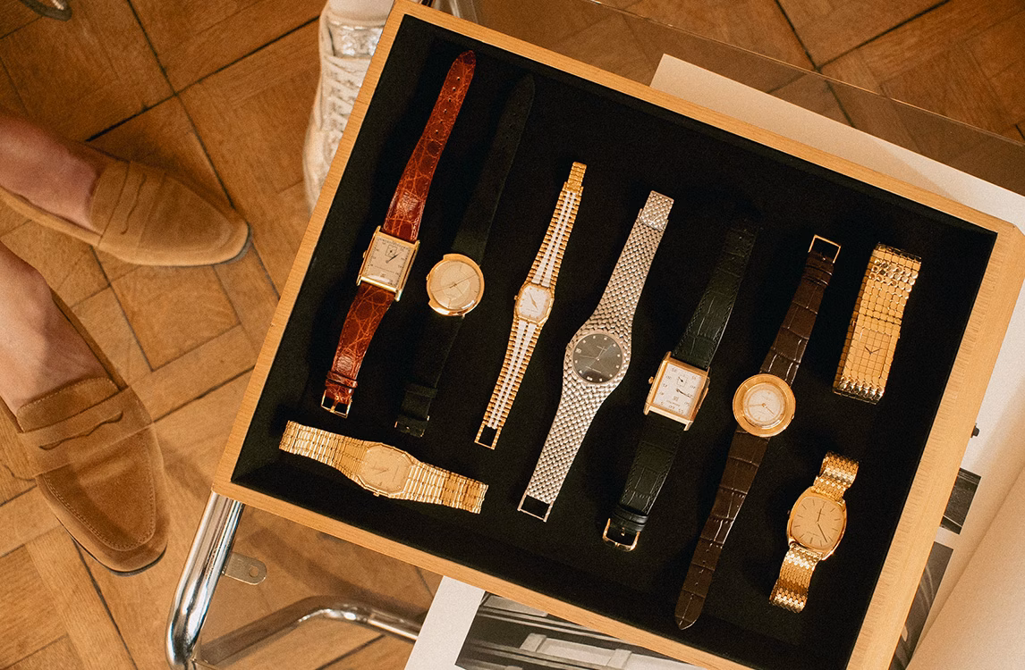 Overhead view of a wooden tray holding ten assorted vintage watches, next to leather shoes.
