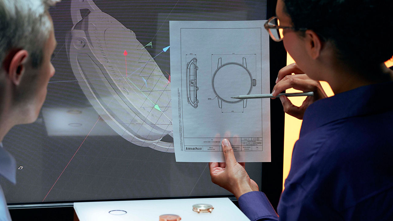 Two colleagues in front of a large screen displaying a 3-D rendering of a timepiece and a technical drawing, discussing some details.